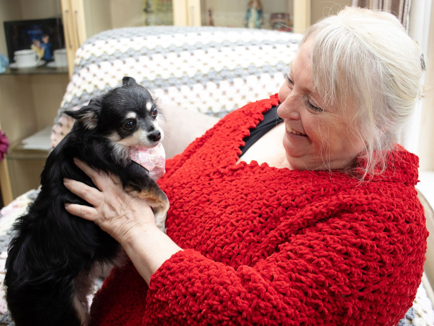 A person in a red sweater holds and smiles at their small black and white dog.