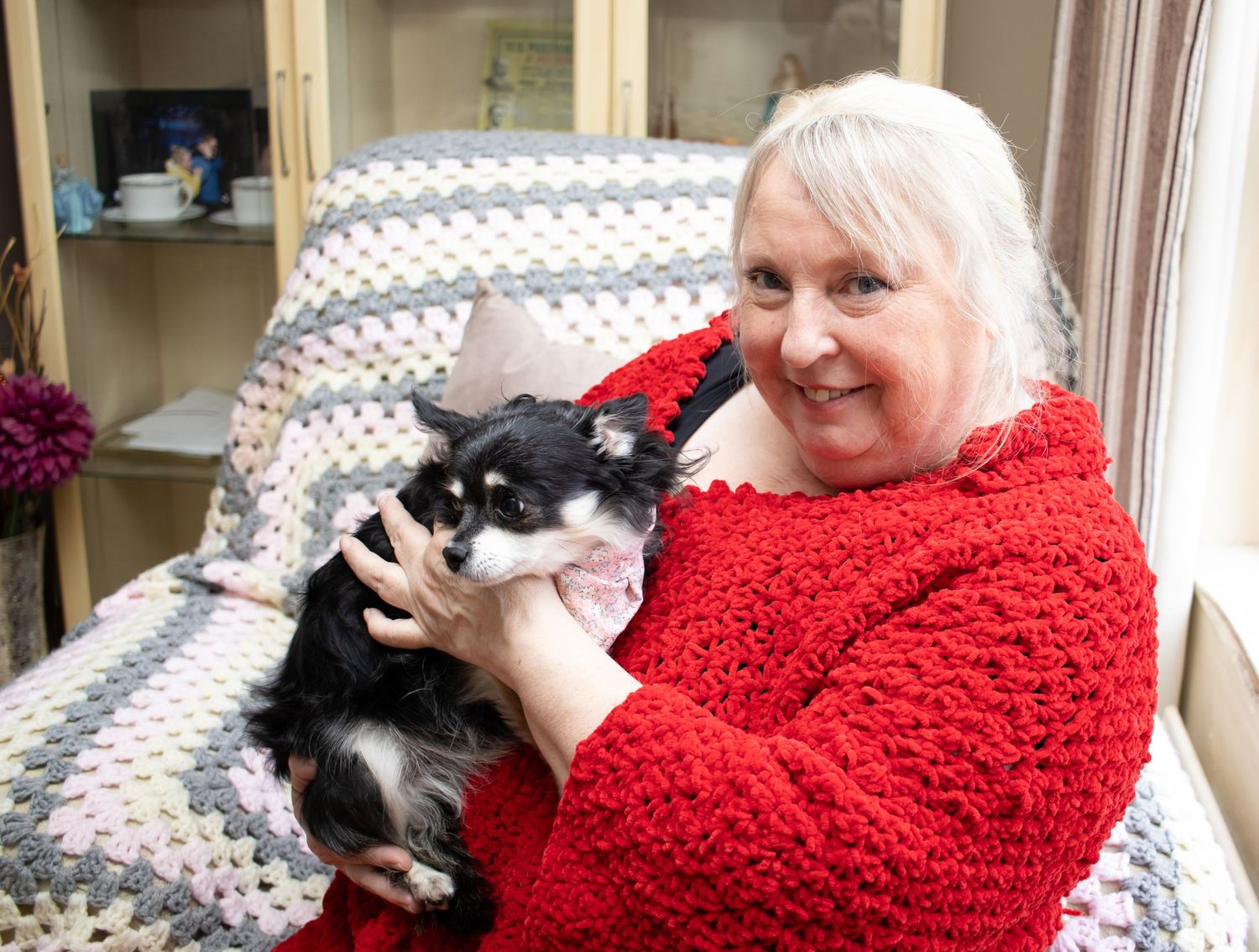 Person in a red sweater sits in a chair cuddling a small black and white dog next to a window.