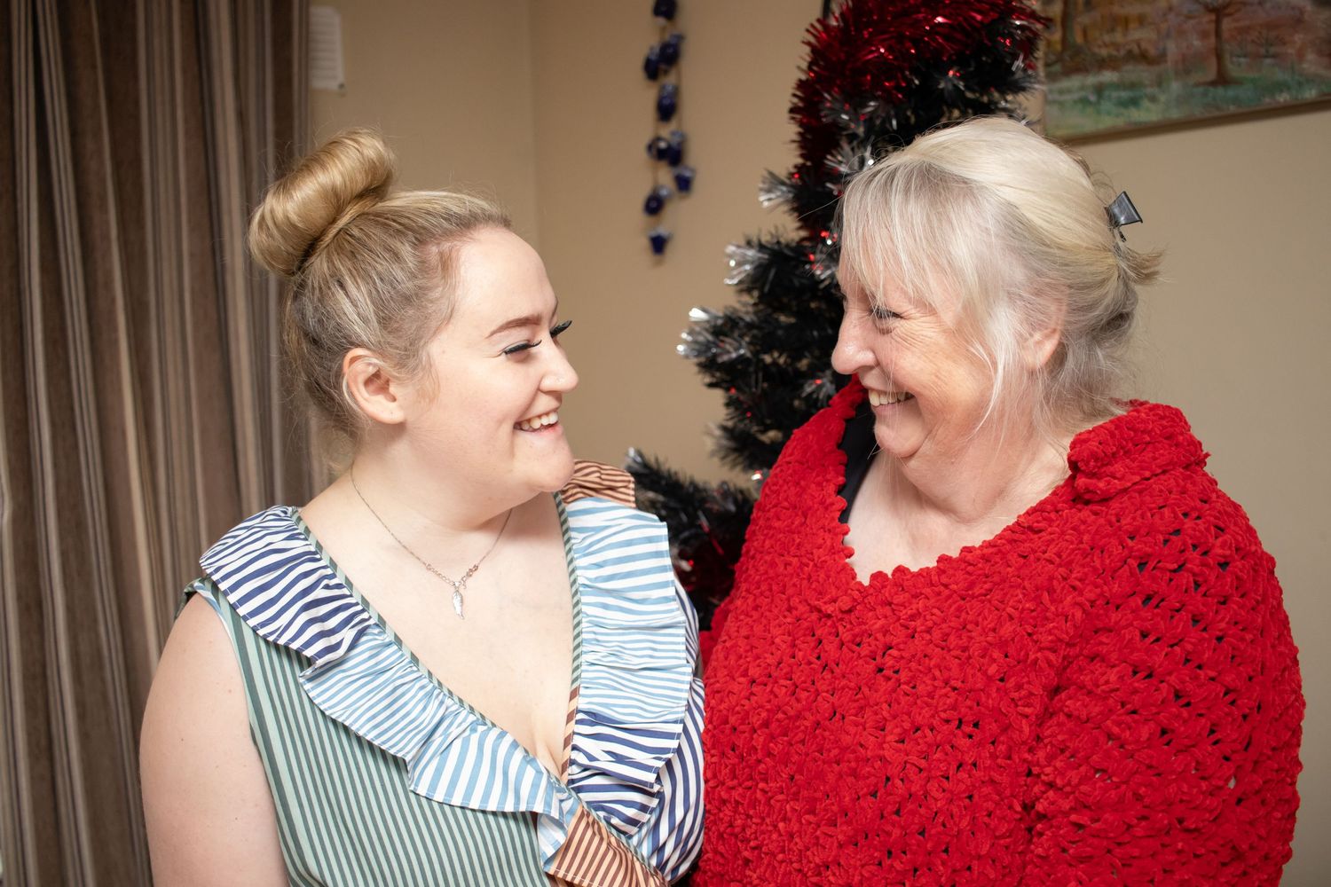 Two people sharing a warm laugh, one wearing a red knit sweater and the other in a ruffled blue dress.