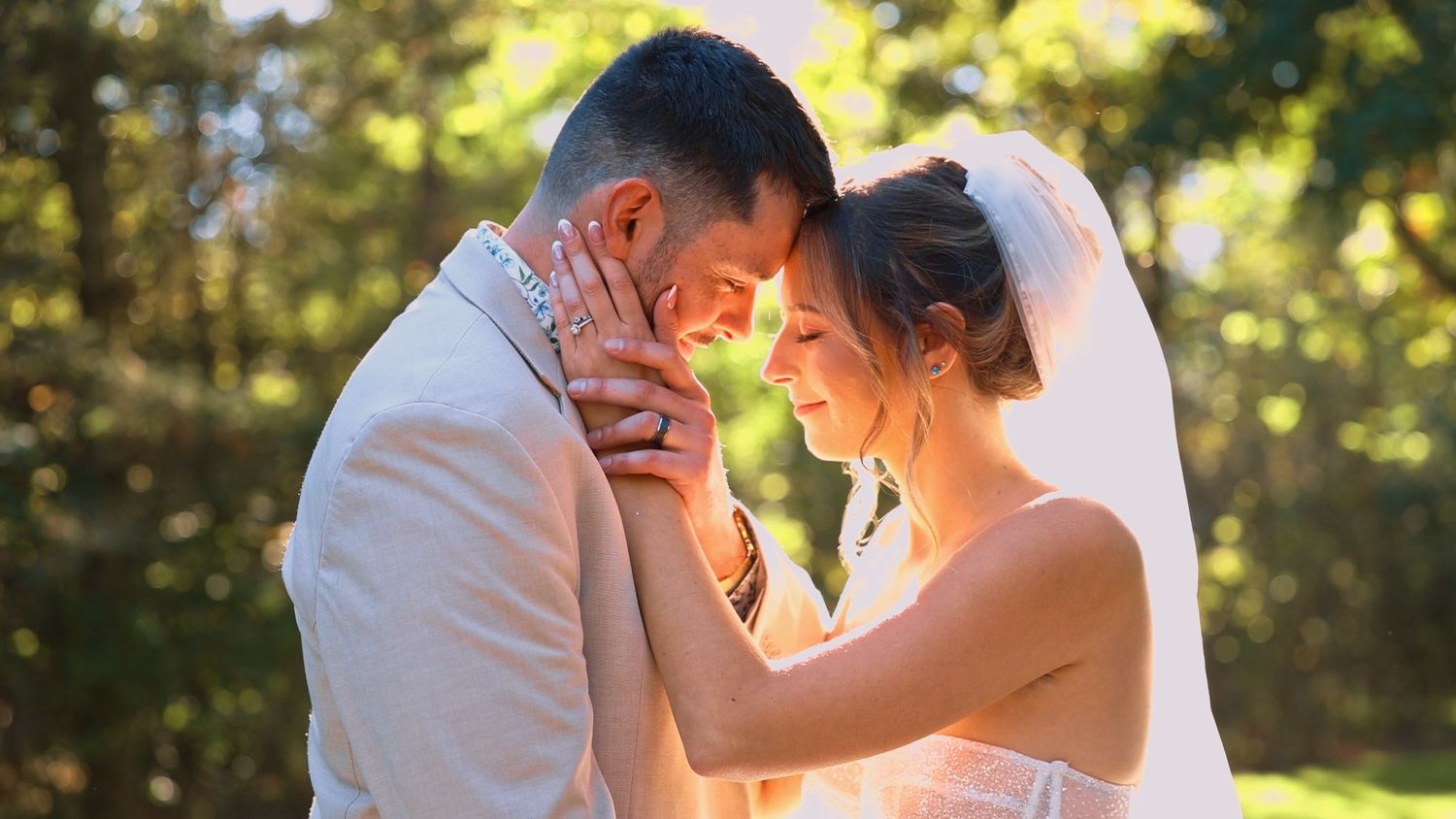 A romantic wedding couple shares an intimate moment in golden sunlight filtering through trees in a forest setting.