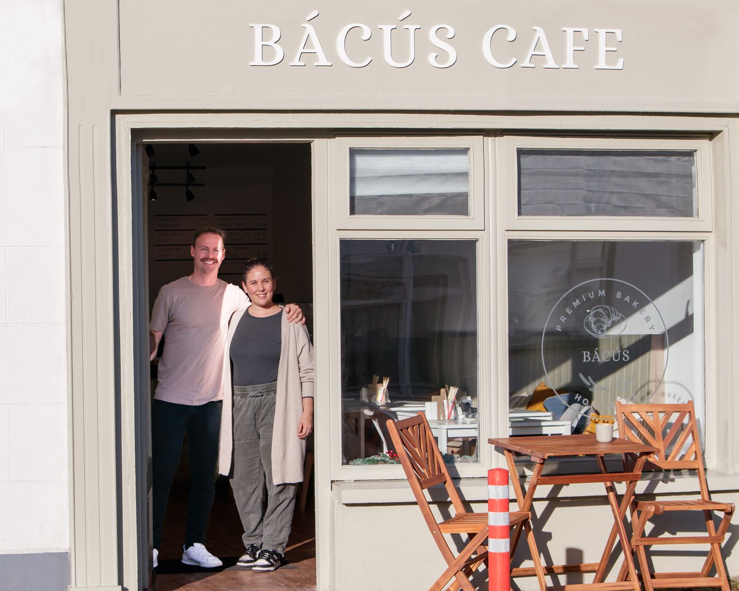 White storefront of Bacus Cafe with wooden outdoor furniture.