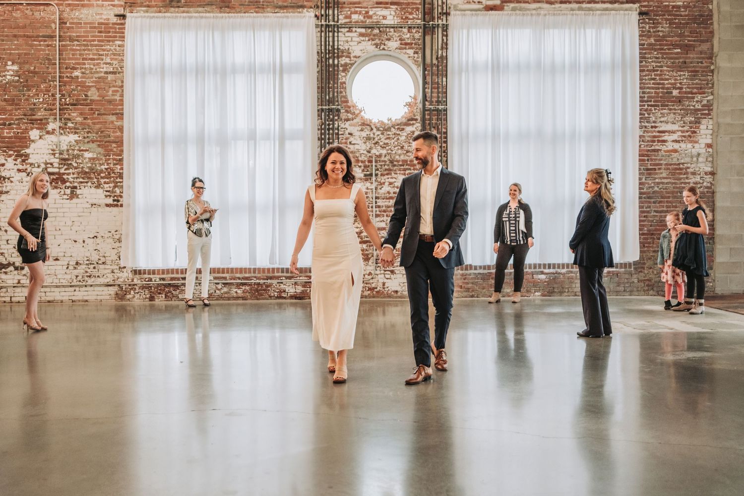 A wedding couple walks together in an industrial venue with exposed brick walls and white curtains as guests look on.  Halo at the Point, ME