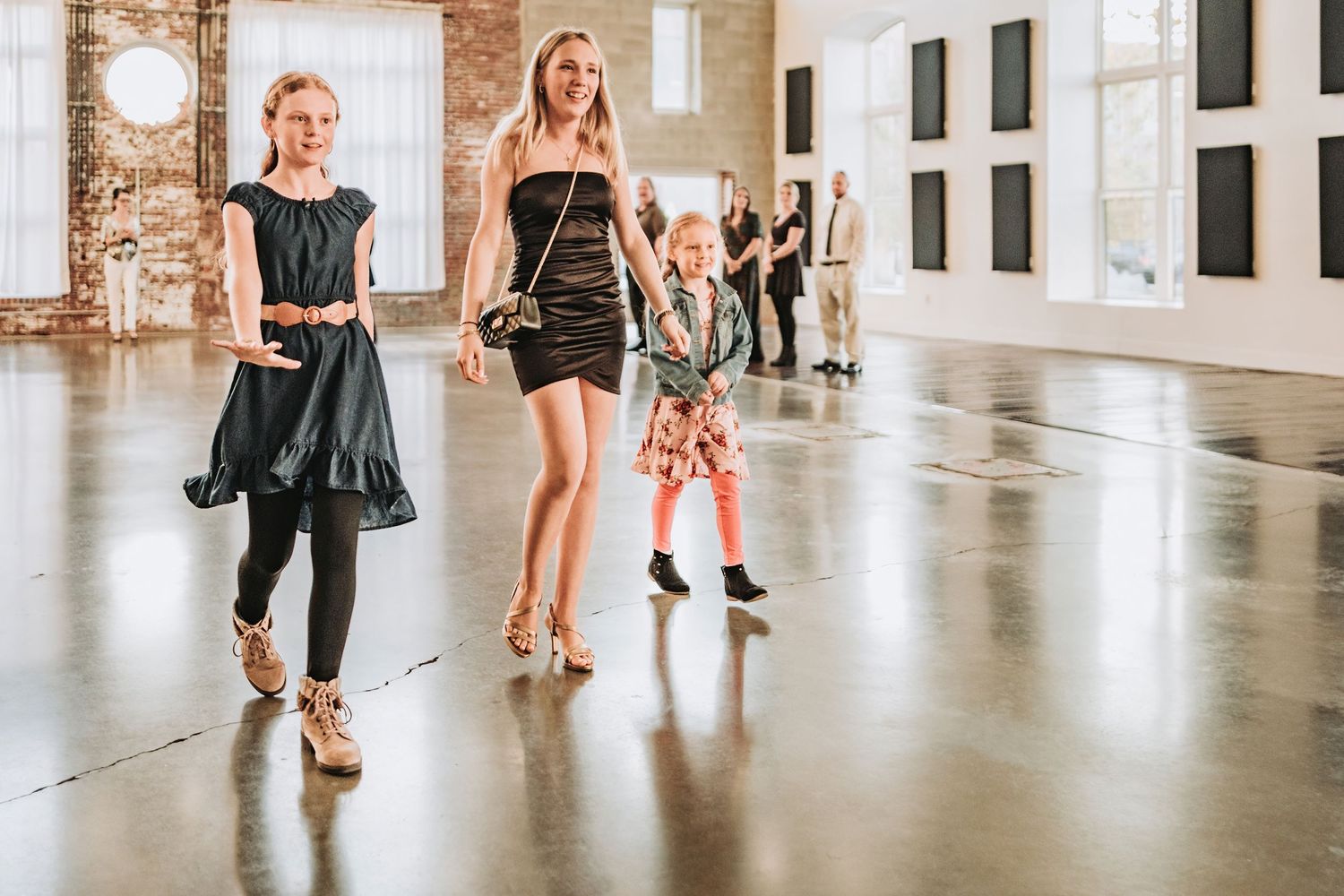 3 young women walk down a sleek industrial hallway wearing elegant evening wear and stylish footwear during a wedding rehearsal.
