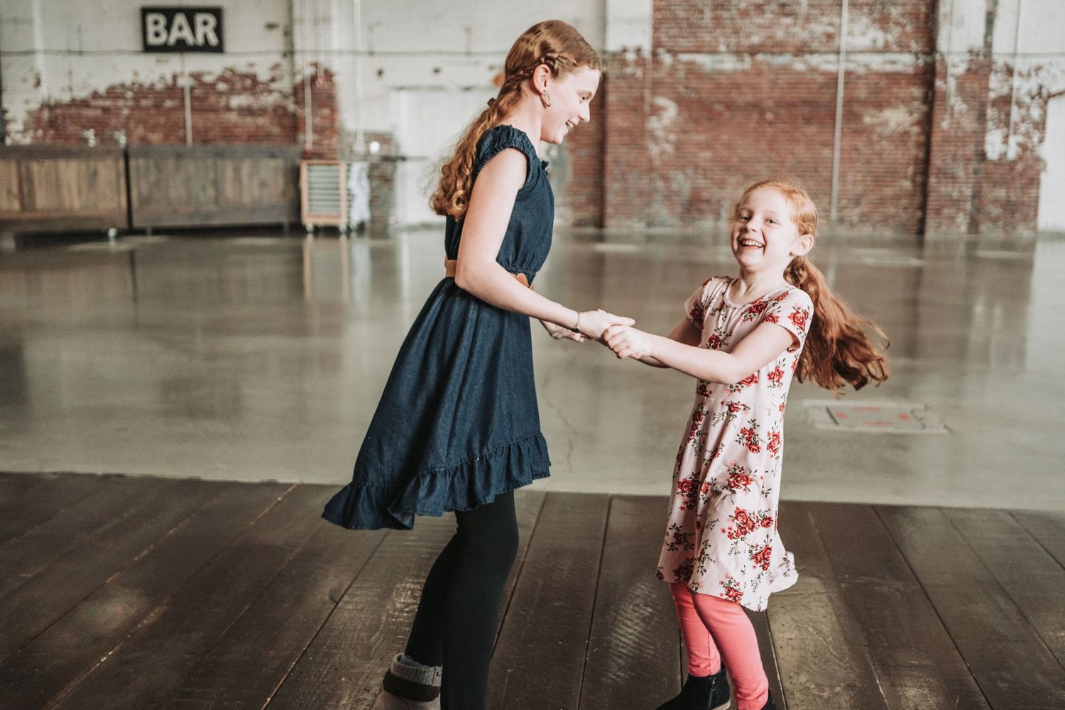 Two sisters dance together playfully on a wooden floor in a spacious room with brick walls and a BAR sign located at Halo at the Point.