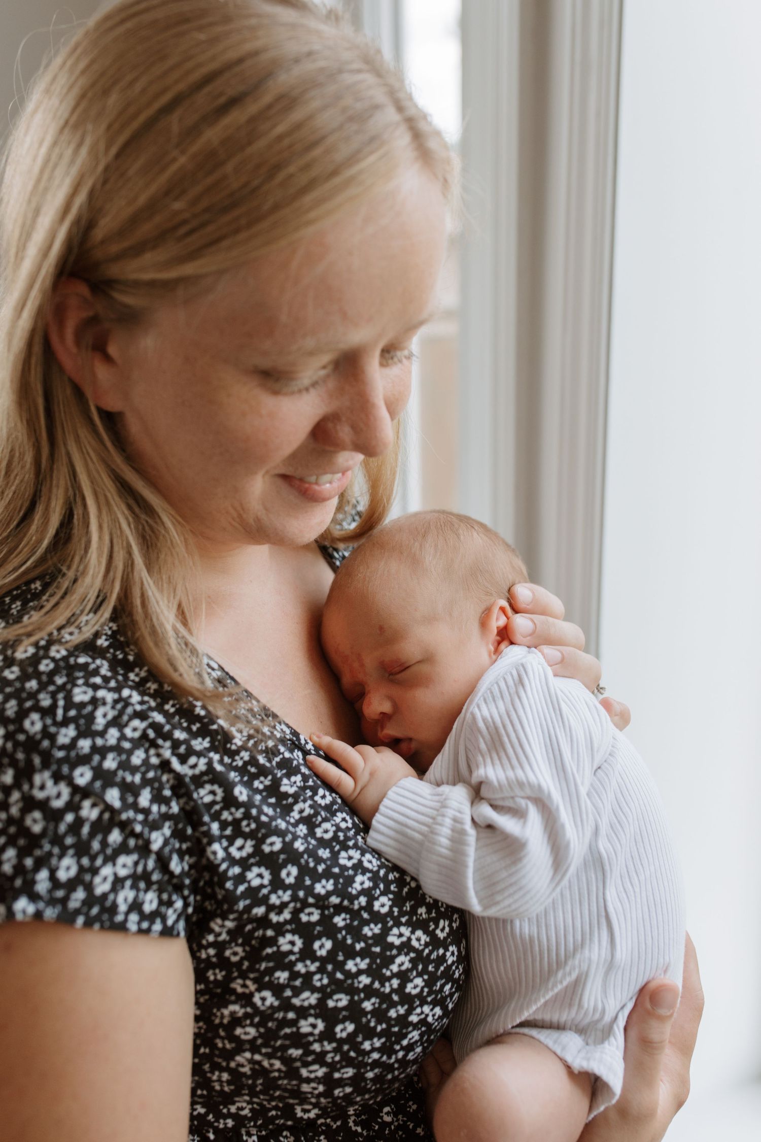 A father in a black polo shirt holds a newborn baby by bright windows.