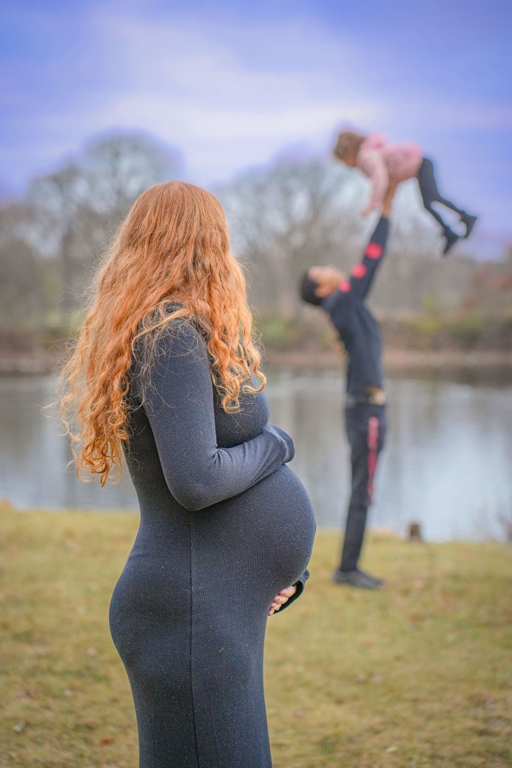 Expectant mother in grey dress stands by lake while child is lifted in background.