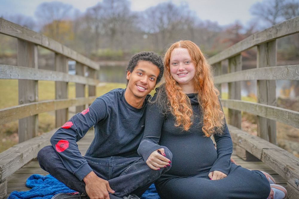 Expecting couple shares intimate moment while sitting on wooden bridge.