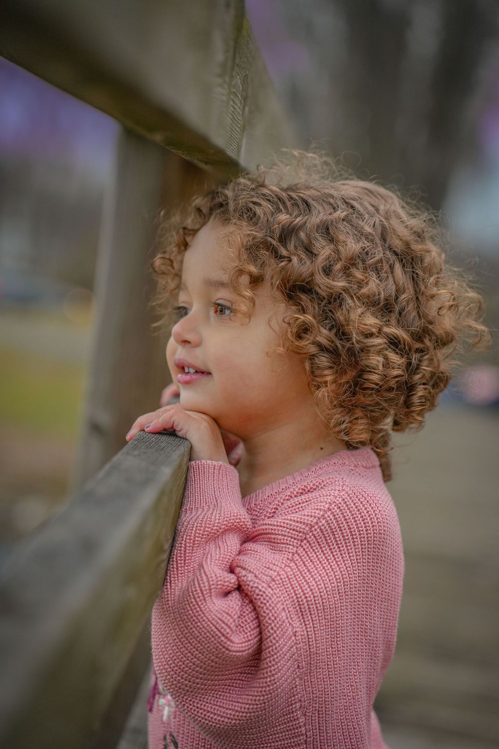 Child in pink sweater leaning on wooden railing with curious expression.