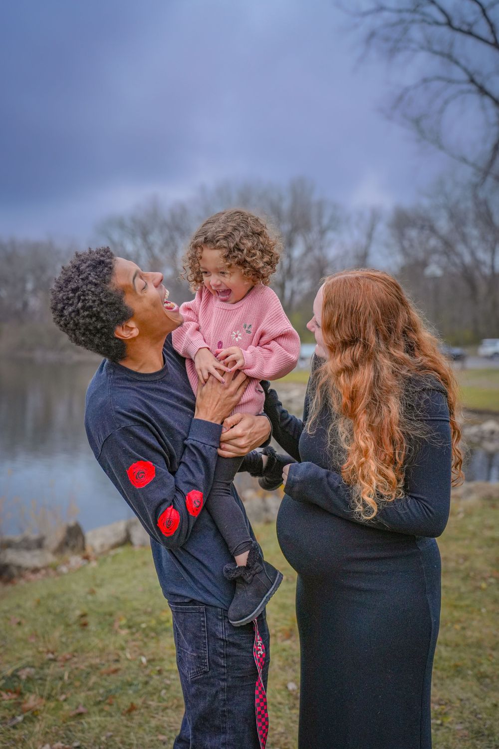 A family shares a joyful moment together by a lake on an overcast day.