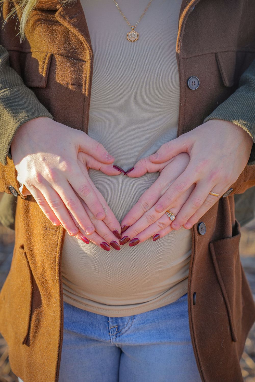 Hands forming a heart shape over a beige top and brown cardigan against a sunny outdoor backdrop.