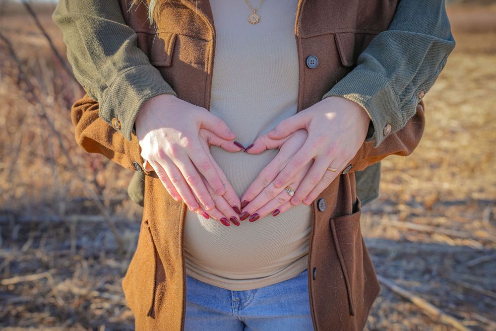 Hands making heart shape over belly while wearing brown jacket and jeans in outdoor setting.