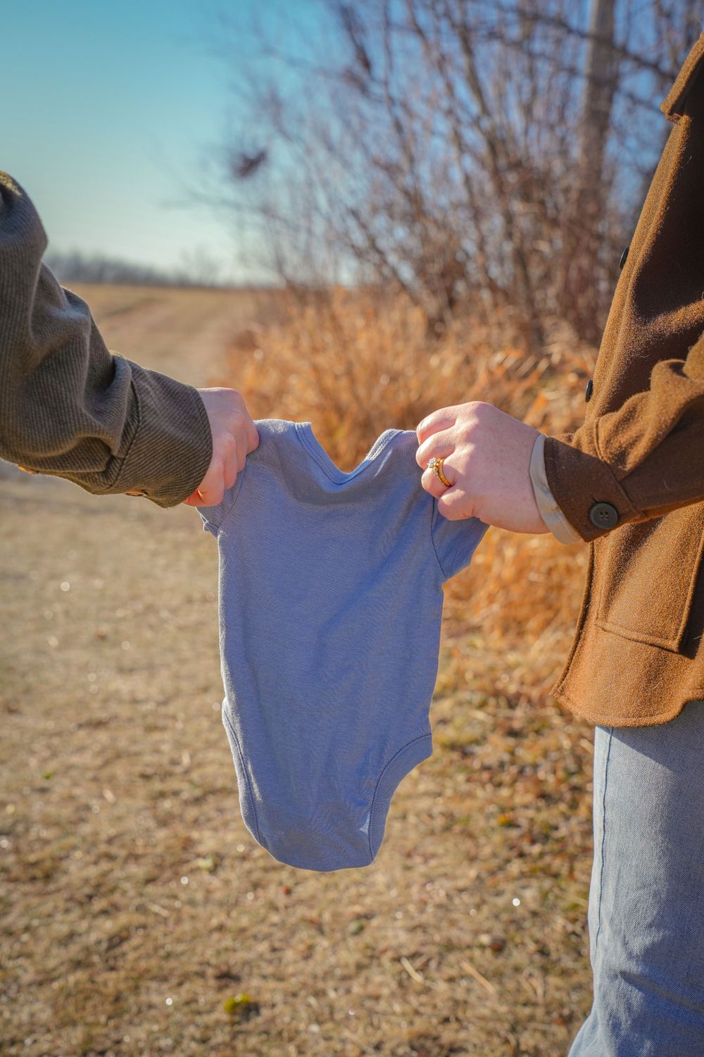 Hands holding a small blue baby onesie against a natural outdoor background.