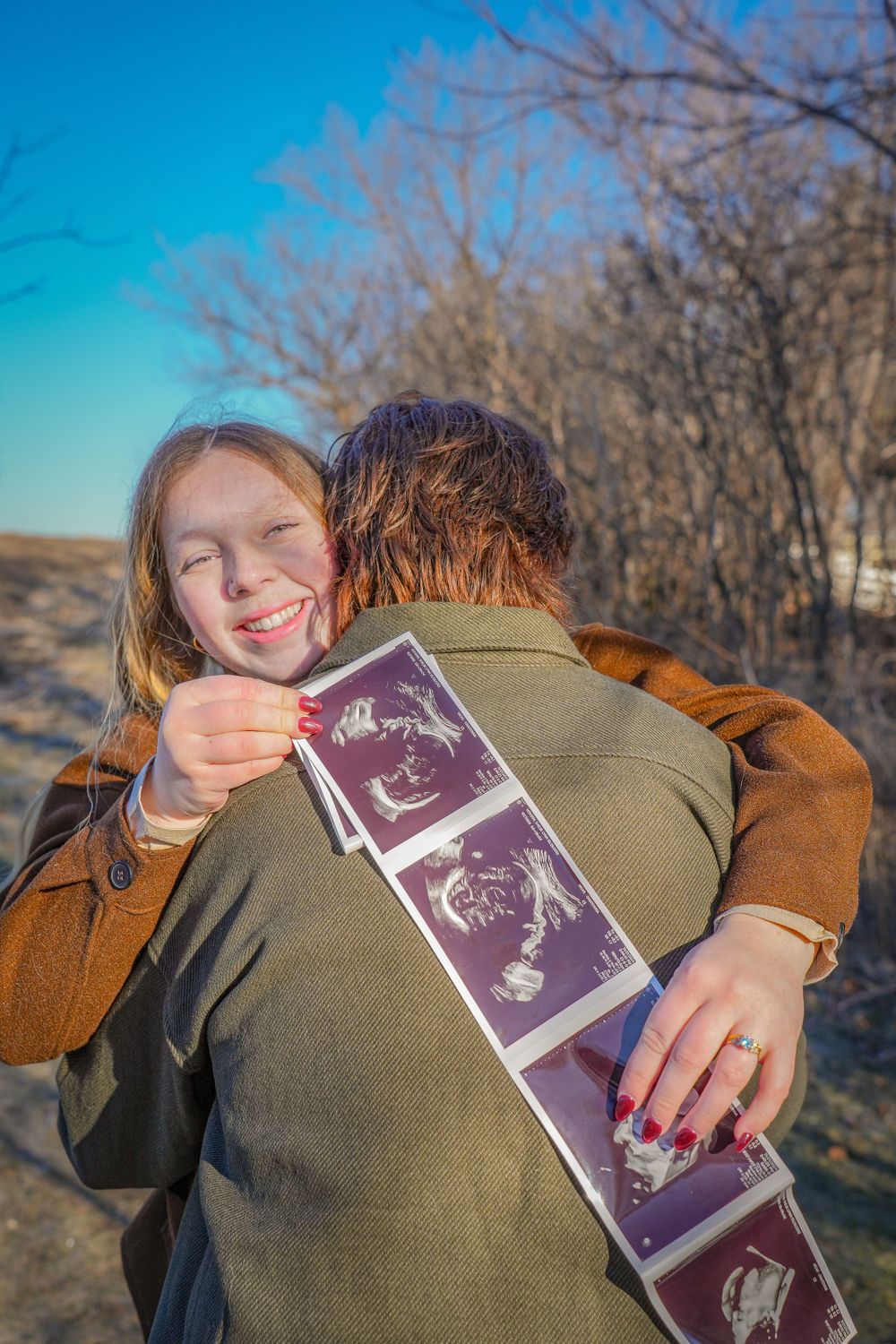 Person holding ultrasound photos while embracing outdoors against blue sky and trees.