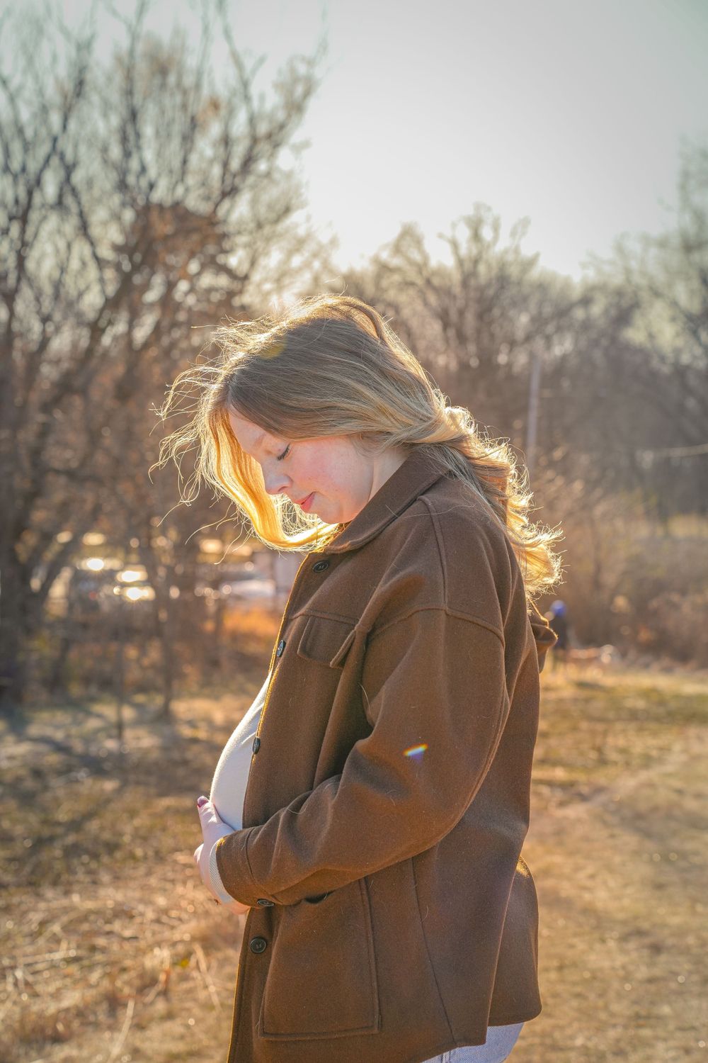 Silhouetted figure in brown coat stands contemplatively in golden sunlight against bare winter trees.