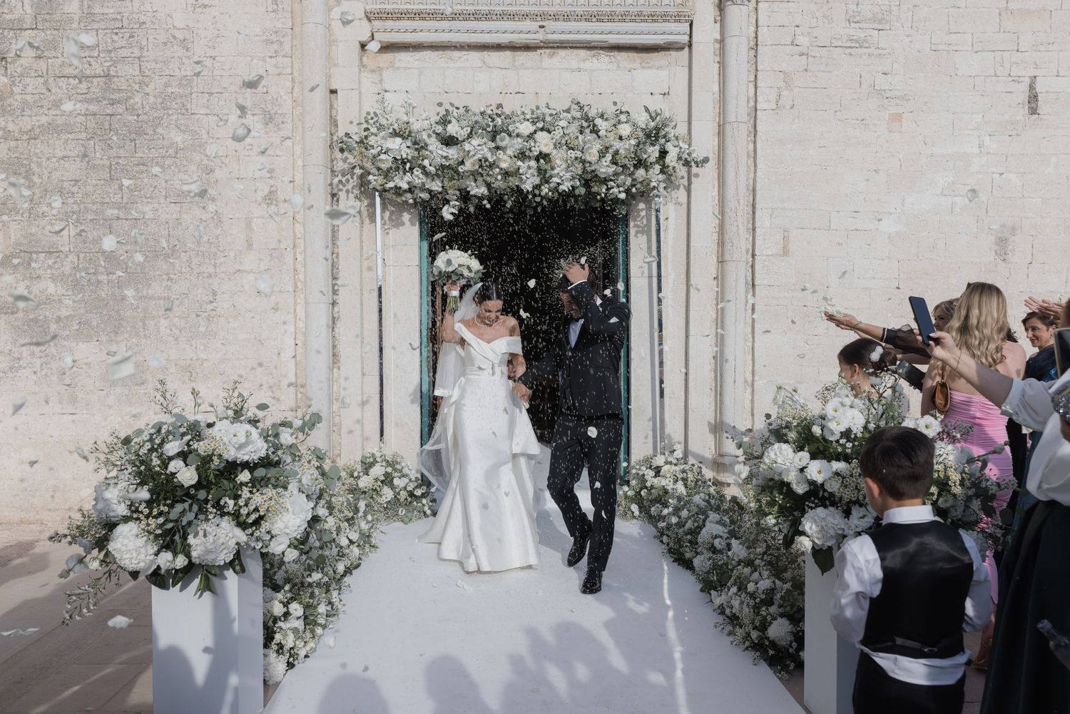 Elegant white wedding ceremony setup with floral arrangements against a brick wall.