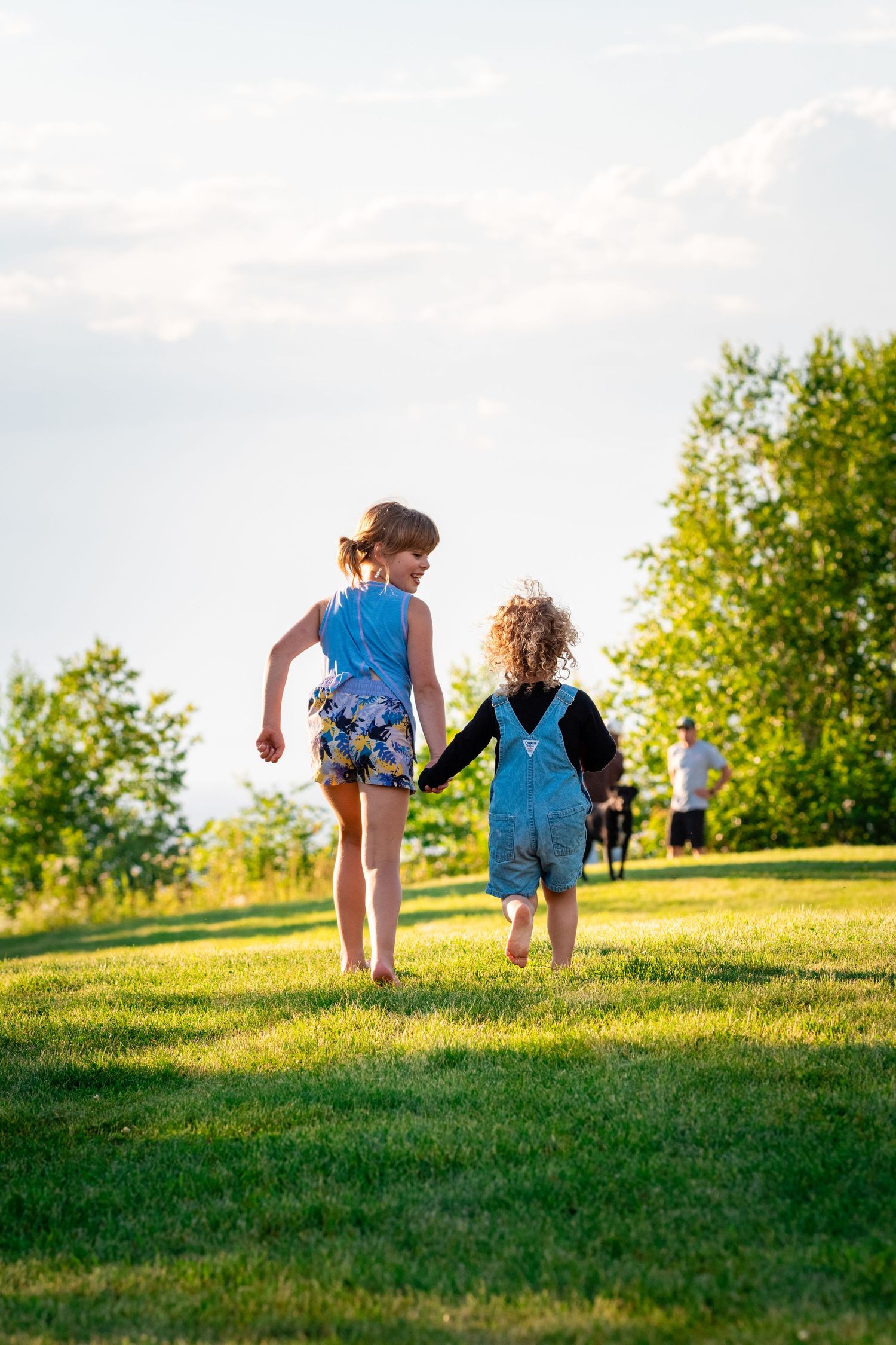 Two children holding hands walking up a grassy hill on a sunny summer day.