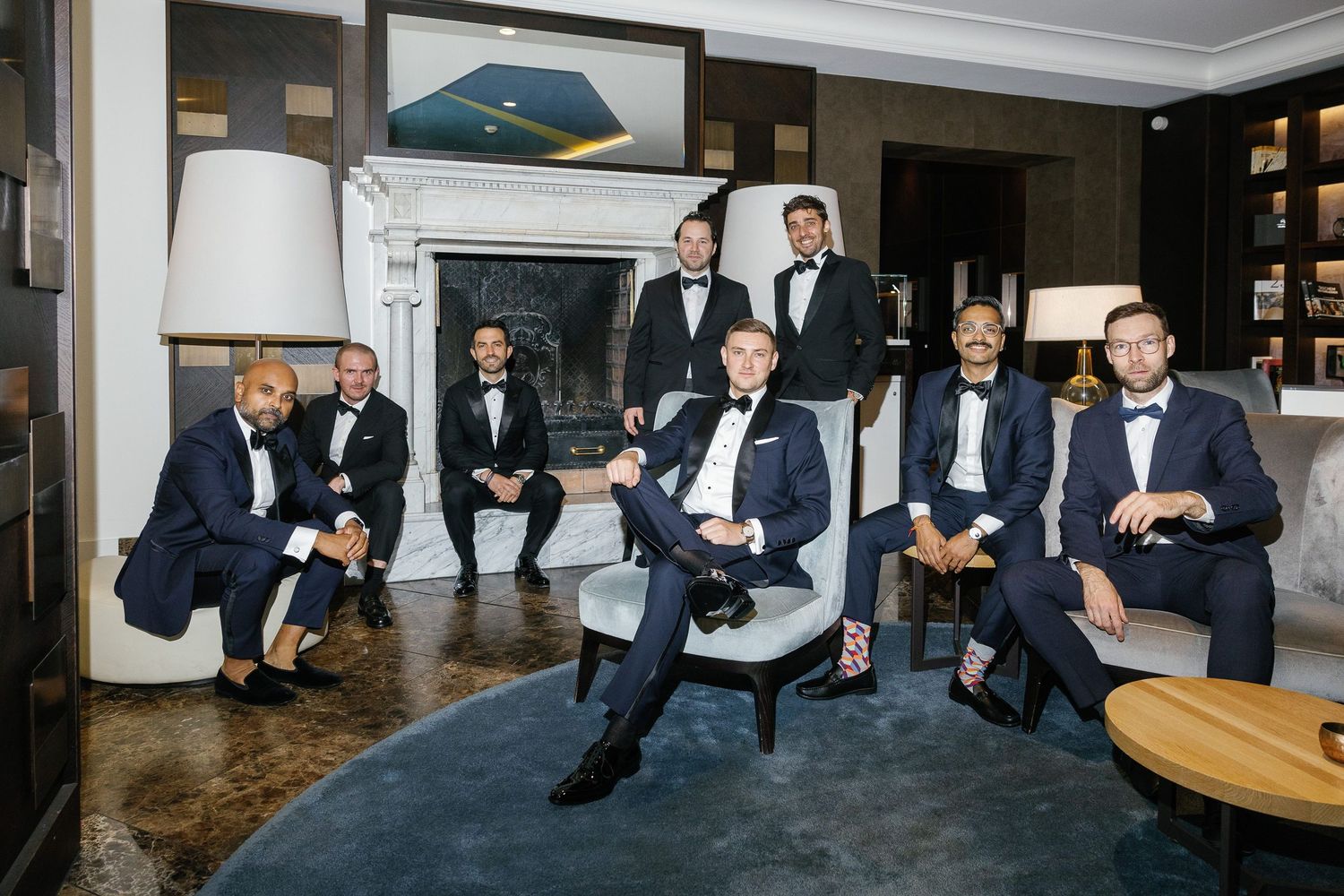 Groomsmen in matching black tuxedos lounging in an elegant room with modern decor and marble flooring.