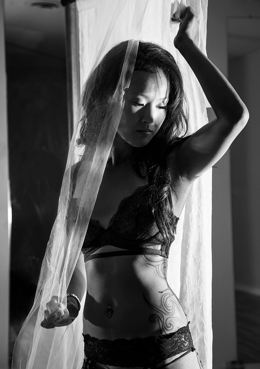 Black and white artistic boudoir photo of figure in lingerie behind sheer curtains in dramatic lighting.