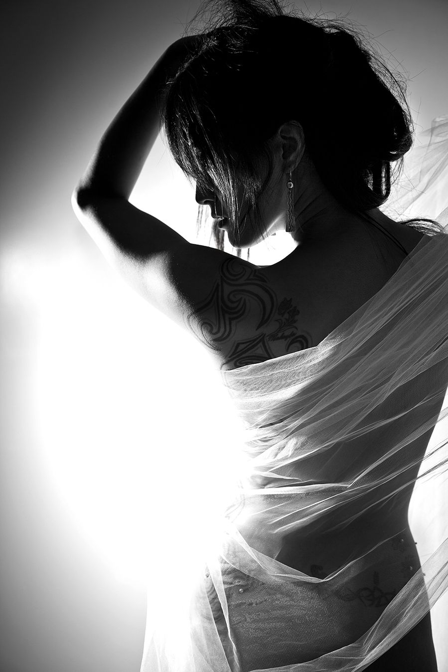 Dramatic black and white silhouette of a person in delicate flowing fabric with a visible shoulder tattoo in backlight.