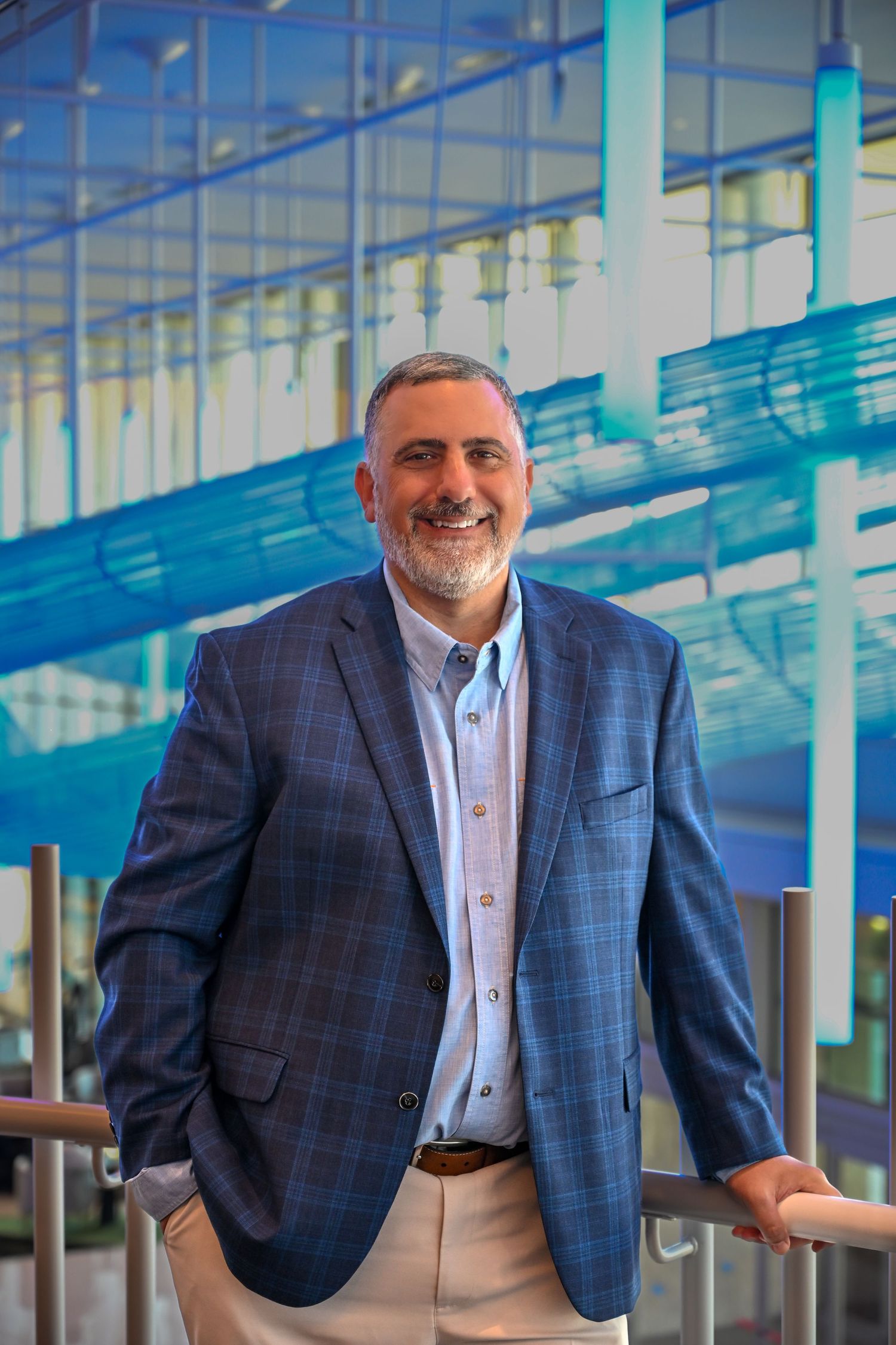 A business executive in a plaid blazer smiles while standing in a modern corporate environment.