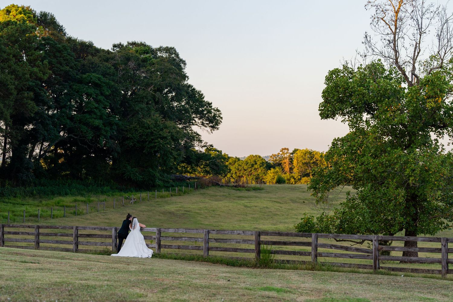 A couple in formal attire stands by a wooden fence at sunset in a pastoral countryside setting.