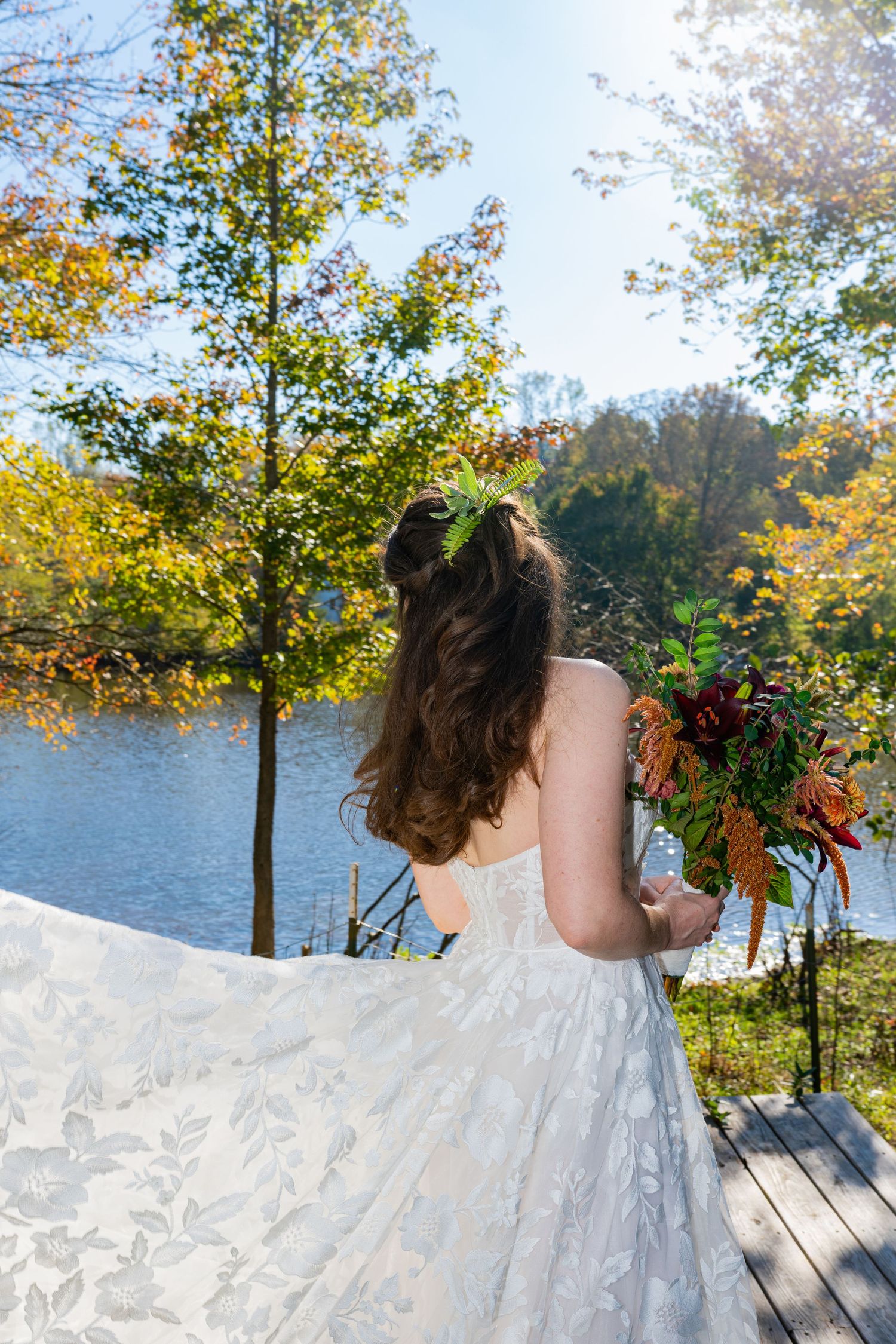 A bride in a white lace dress holds a colorful autumn bouquet while standing on a dock by a lake surrounded by fall foliage.