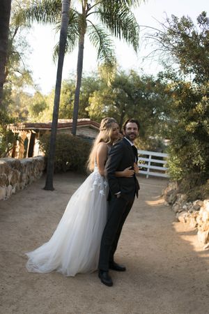 A couple in wedding attire pose together on a dirt path lined with palm trees and stone walls at an outdoor venue.