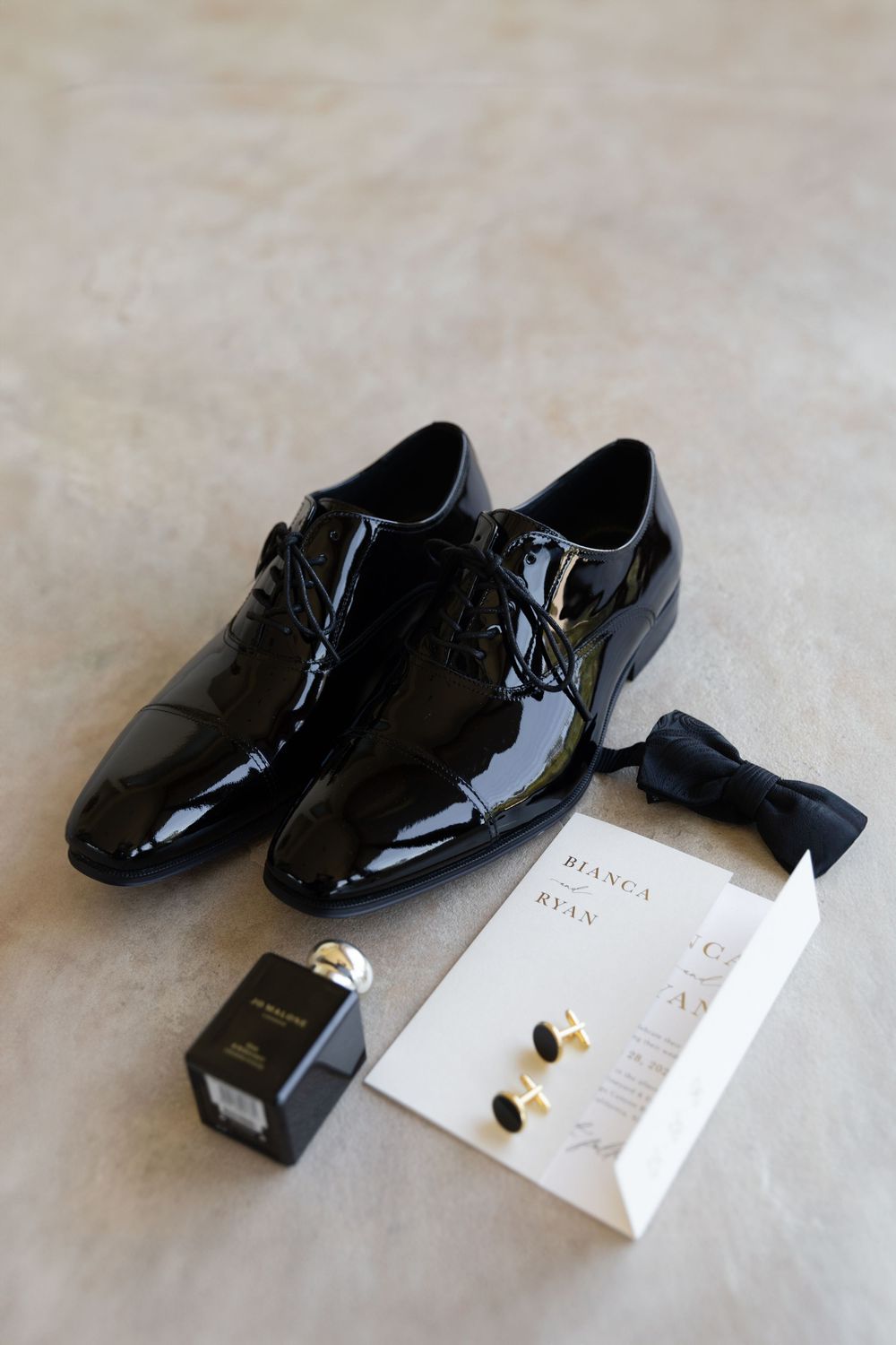 A pair of shiny black patent leather dress shoes with laces displayed next to white cards on a light gray surface.