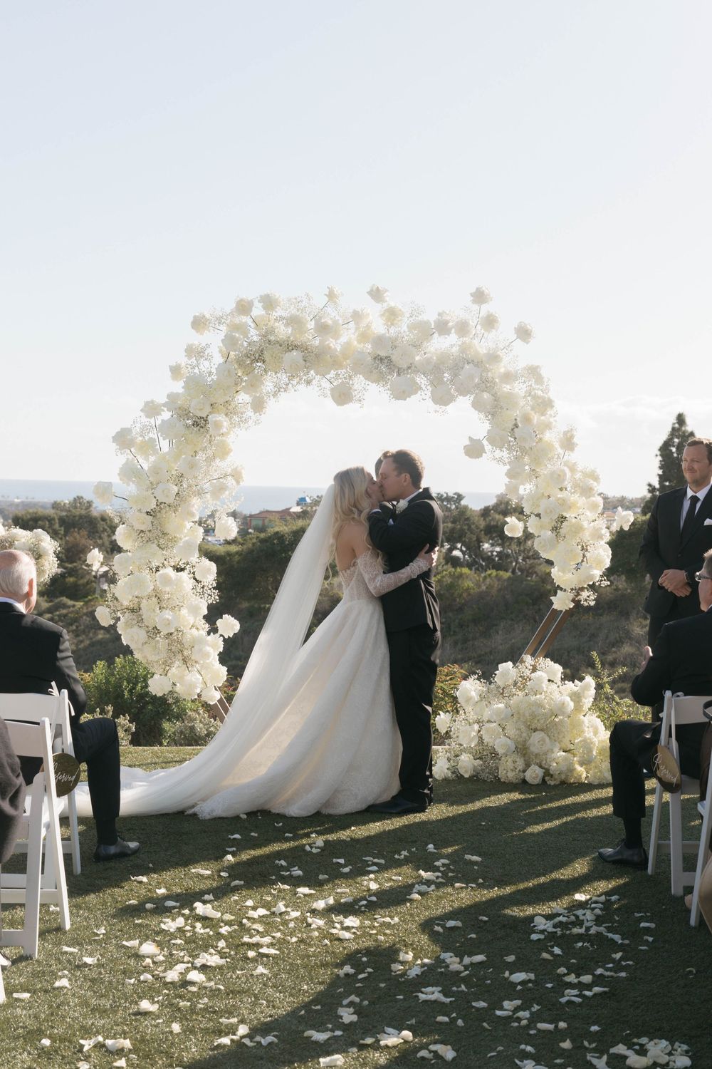 Outdoor wedding ceremony setup with white floral arches and scattered petals along a grassy hillside at sunset.