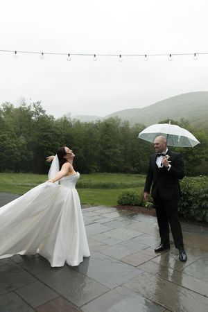 A couple poses with umbrellas in the rain during their wedding photoshoot on a stone patio with green hills behind them.