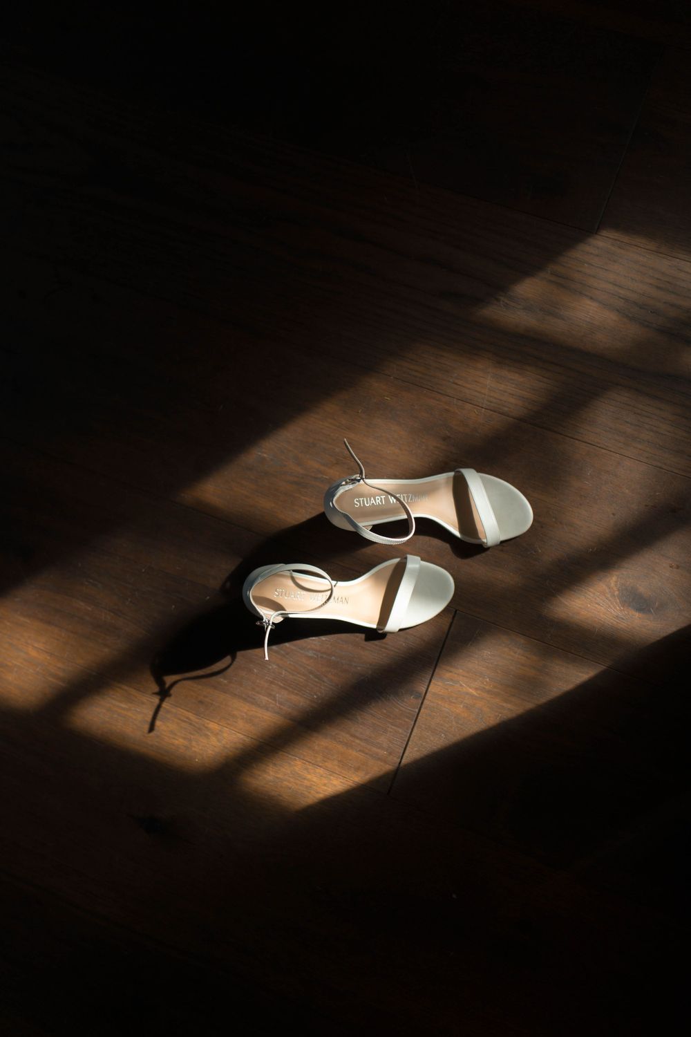 White wedding sandals casting dramatic shadows on a dark wooden floor.