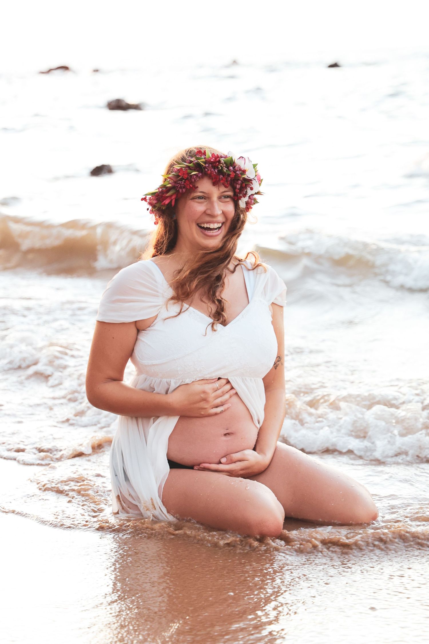 An expectant mother wearing a flower crown sits in the ocean waves during a maternity photoshoot.