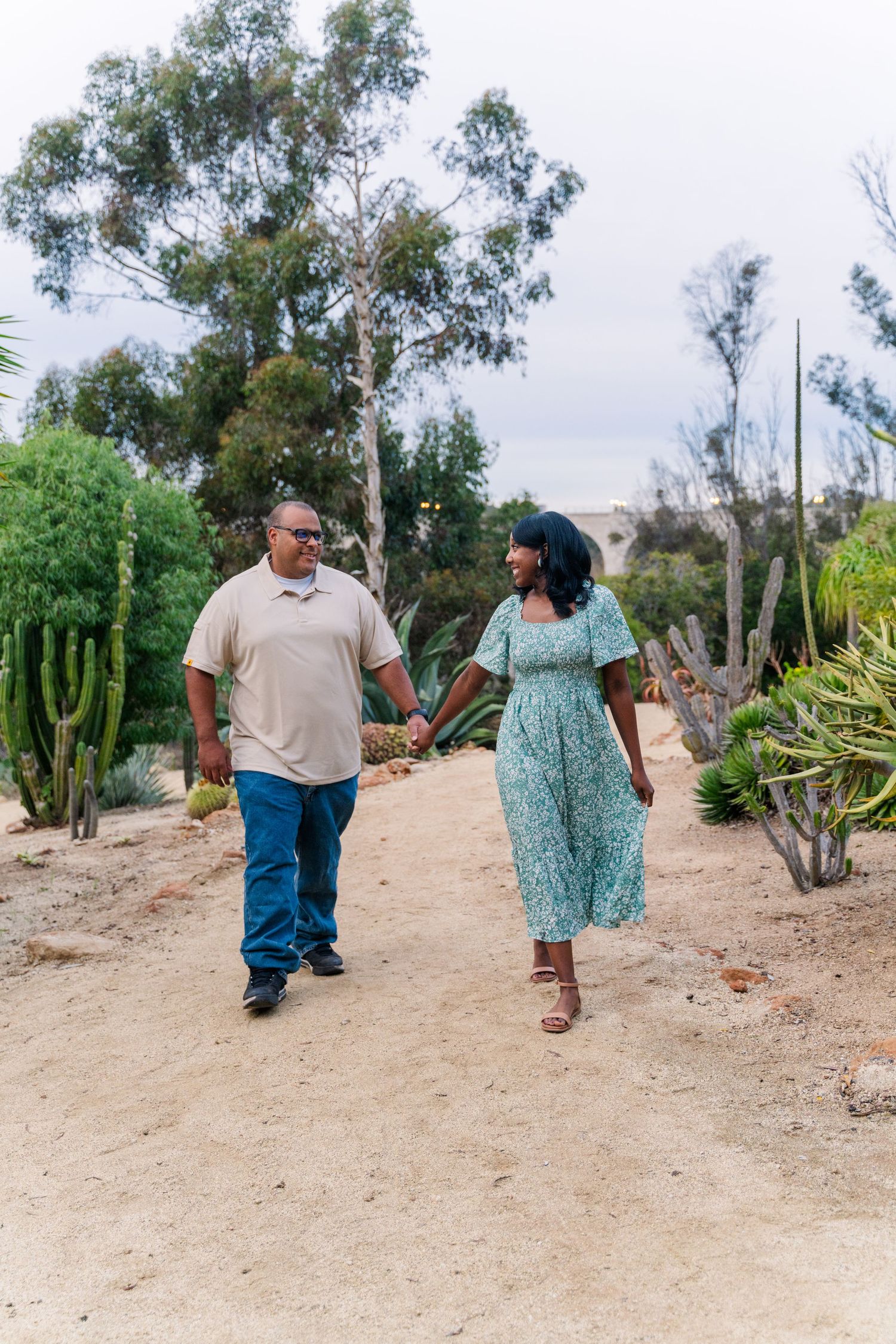 A couple walks hand in hand along a desert garden path with tall cacti and eucalyptus trees in the background.