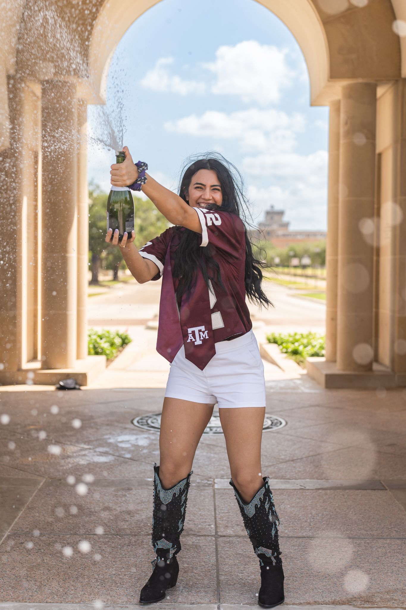 Texas A&M graduate popping champagne while wearing cowboy boots and a maroon stole at the Albritton Bell Tower in College Station.