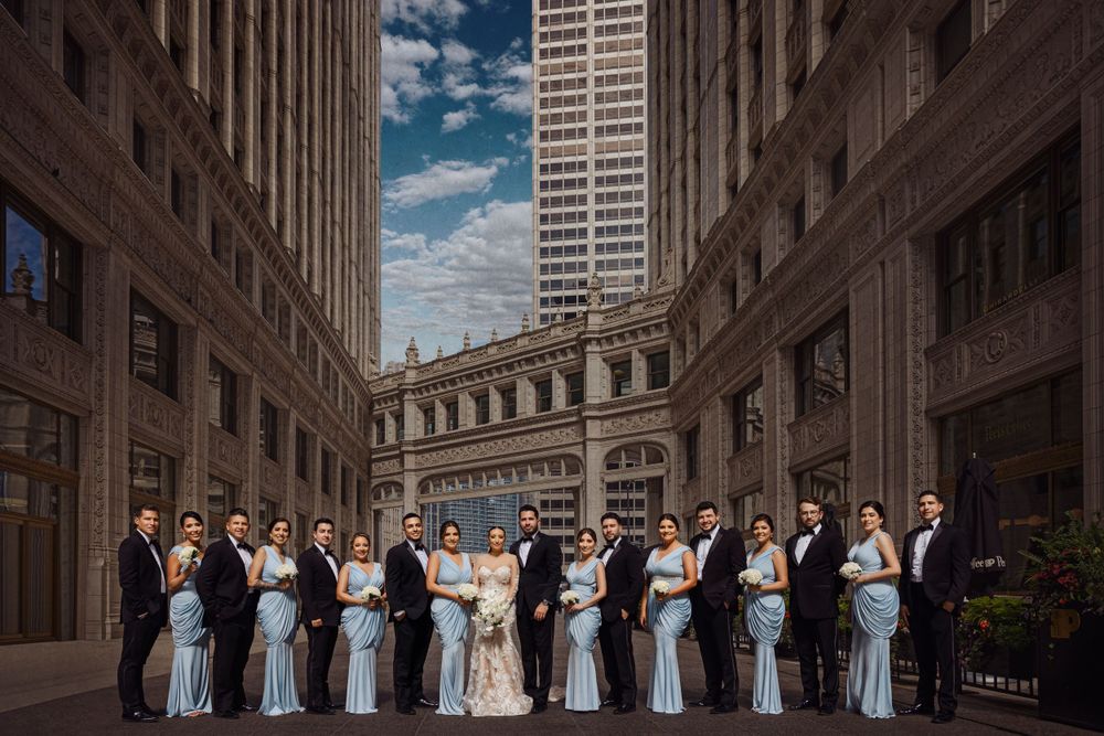 Wedding party poses in an urban setting between tall buildings under a blue sky.