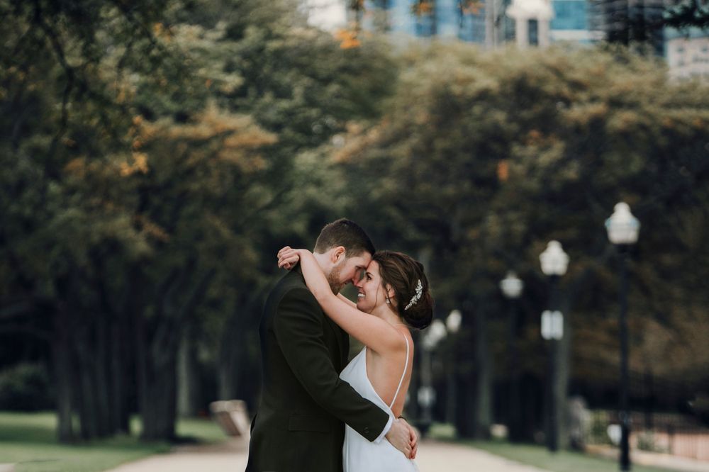 A bride and groom share an intimate embrace in a park setting with trees and lampposts in the background.