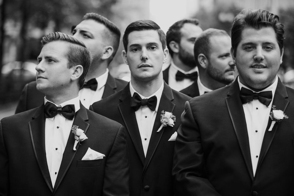Groomsmen in black tuxedos and bow ties stand together looking off to the side in a formal wedding portrait.