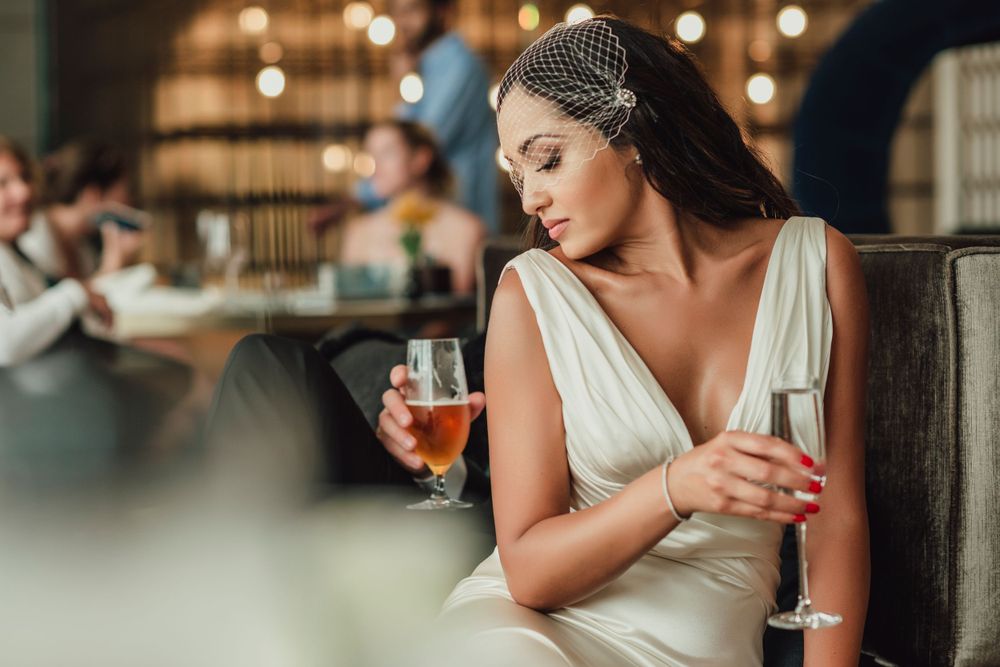Elegant bride in a white dress and headpiece sits at a bar holding a cocktail glass.