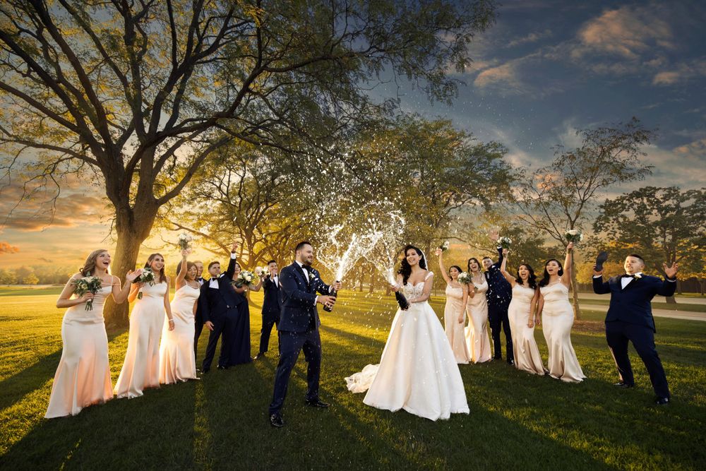 Wedding party celebrates on a grassy field during golden sunset with a large tree in the background.
