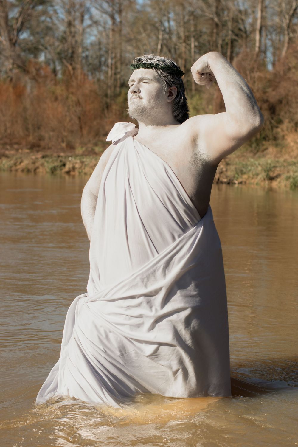 A person wearing a white toga-like garment poses dramatically in water with one arm raised against an autumn riverside backdrop.