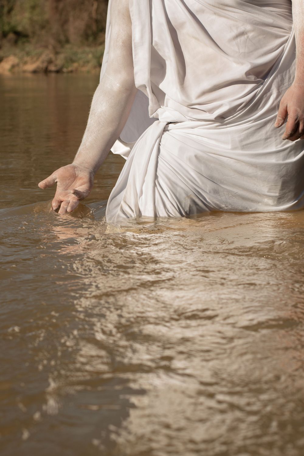 Person wearing flowy white fabric stands in shallow river water, reaching down to touch the surface.