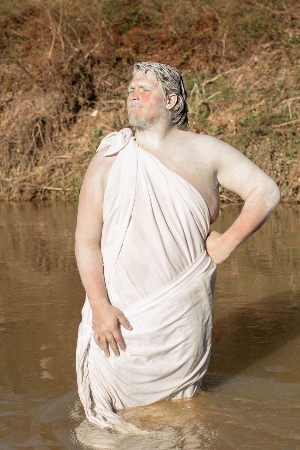Person wearing white toga-style garment stands in shallow water with dramatic pose against natural backdrop.