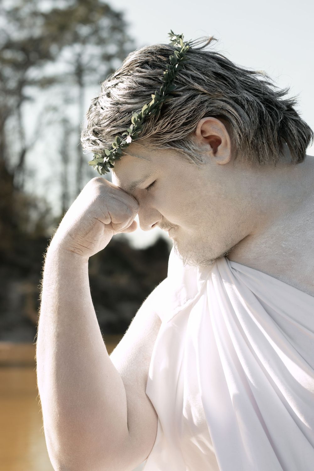 Figure in white clothing wearing small flowers in their hair bows head in contemplative pose against sunlit forest backdrop.