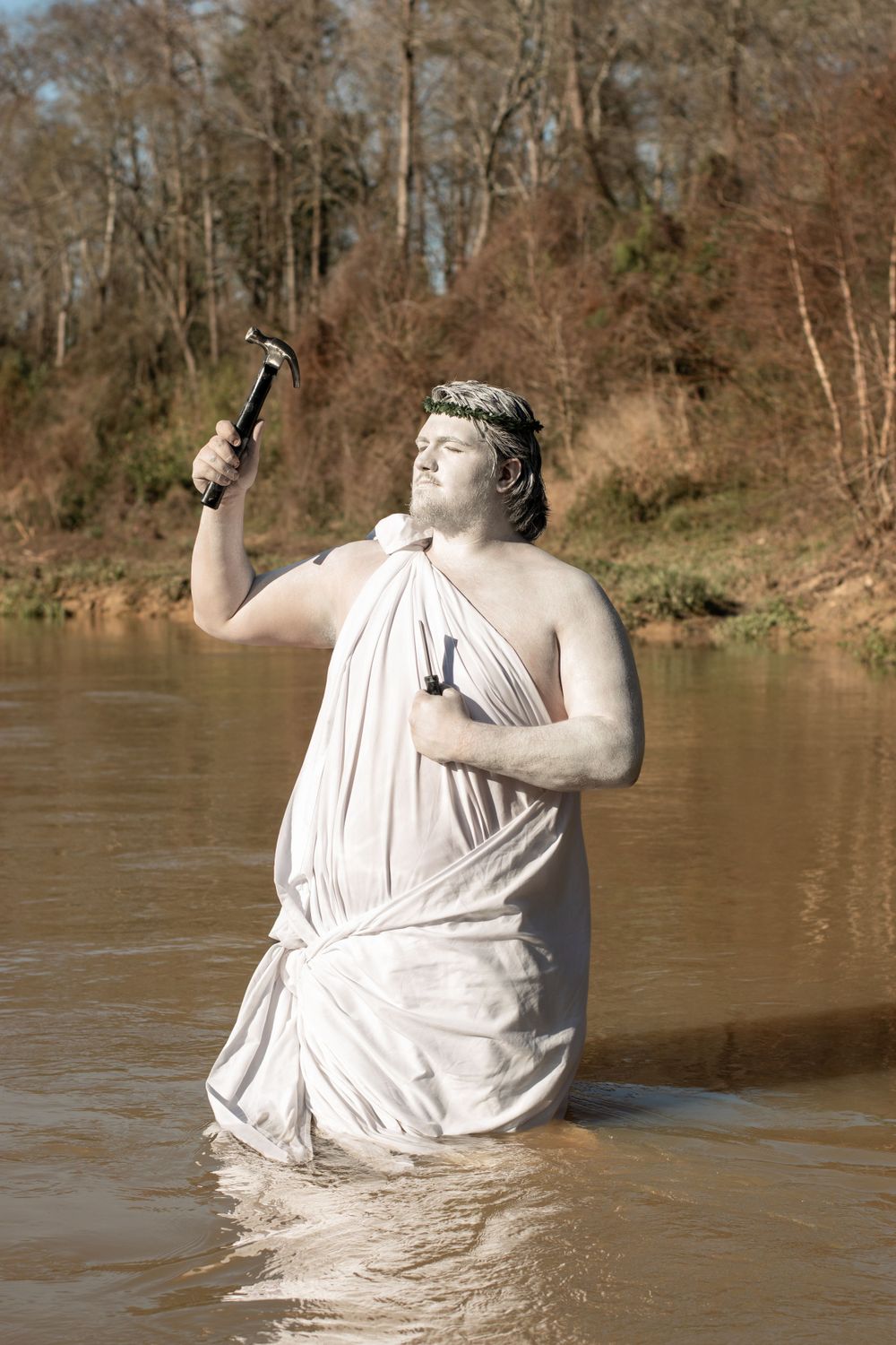 Figure in draped white cloth stands waist-deep in river water while holding a bottle aloft.