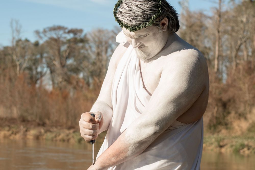 A person in a light-colored shirt stands near a river on a sunny day with autumn trees in the background.