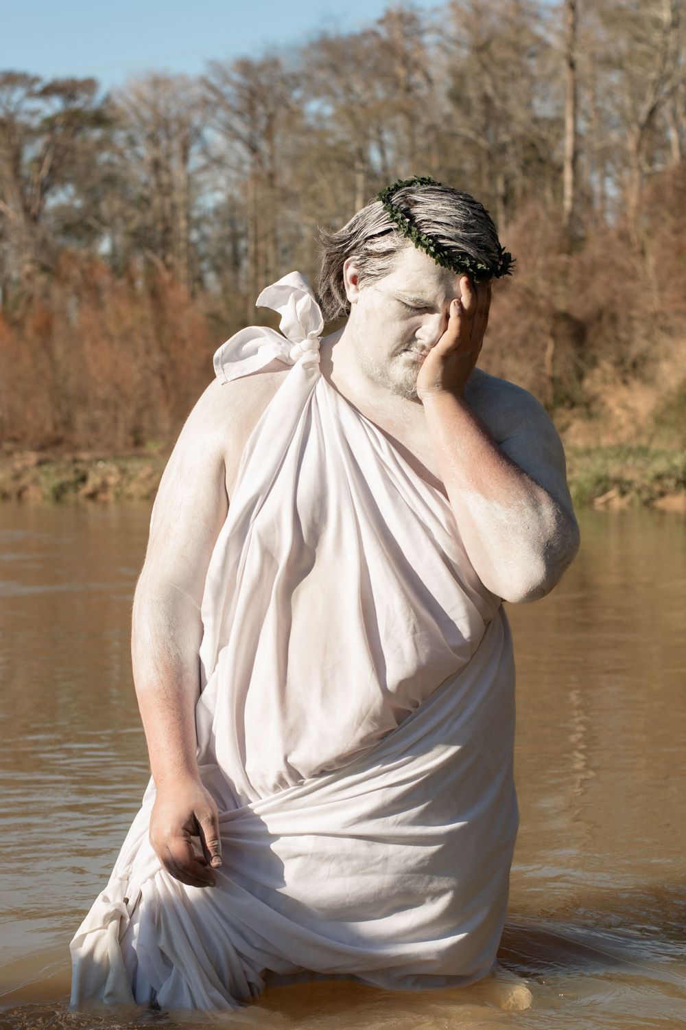 A person wearing a white draped toga-style dress poses in water with a dramatic gesture near trees by the riverbank.