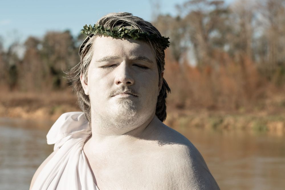 A person wearing a light-colored garment poses with closed eyes in front of a natural water setting on a sunny day.
