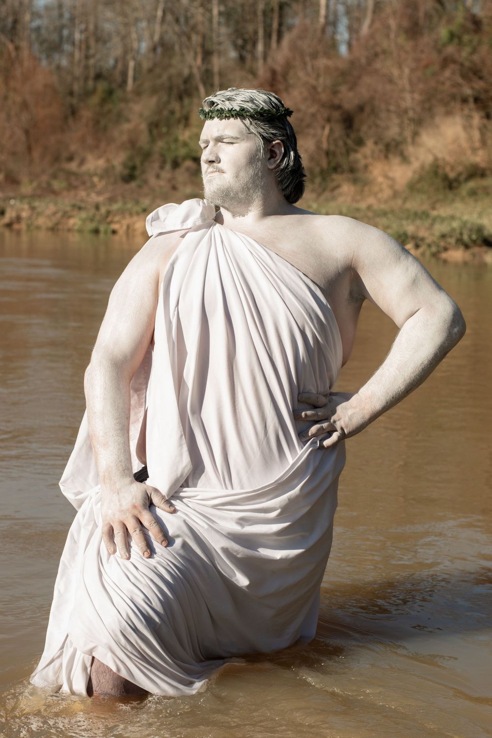 Person posed in classical Greco-Roman style wearing white toga stands in shallow water outdoors with brown landscape.