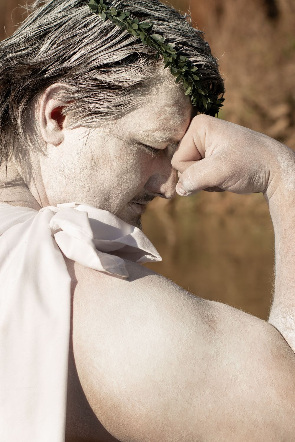 Figure wearing a laurel wreath crown and light clothing presses hand to face in contemplative pose against blurred background.