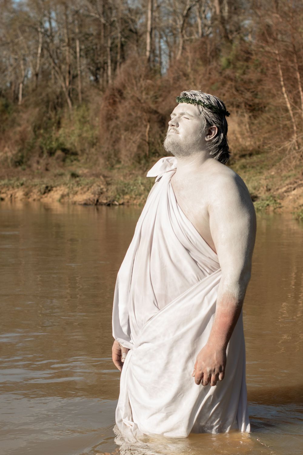 A person wearing a white toga-style garment stands in water while tilting their head back toward the sky.